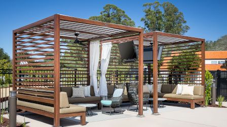 A modern outdoor patio area with wooden pergolas, seating, tables, and a TV. Surrounded by trees and landscaped greenery, under a clear sky.