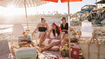 Four friends lounging on a sunny beach with umbrellas, snacks, and vintage suitcases, relaxing and chatting near the shore.