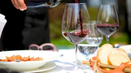 A person pours red wine into a glass at a table with bread and food, outdoors, sunset vibe.