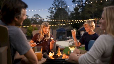 A group of four people sits around a fire pit, enjoying roasted marshmallows and drinks, with string lights overhead and trees in the background.