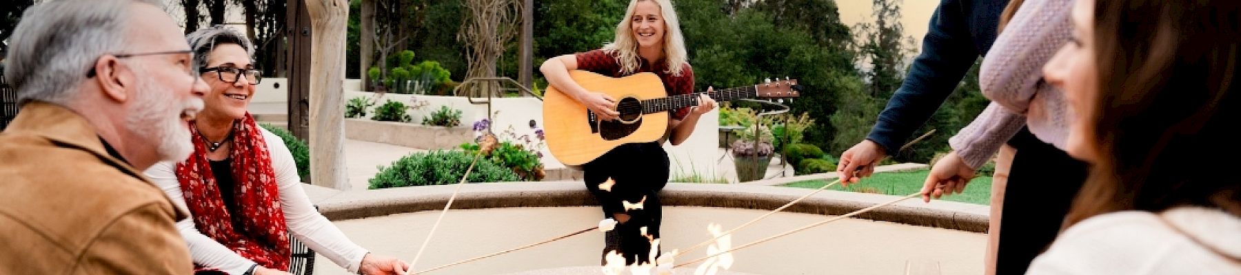 A group of friends sits around a fire pit while one person plays a guitar and sings at sunset.
