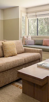 A cozy living room with a beige sofa, a wooden coffee table, a window seat with cushions, a floor lamp, and a warm, mid-century style decor.