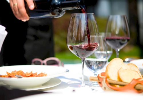 A person pours red wine into a glass at a table set with bread, water, and plates, during a dining scene.