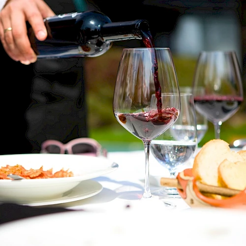 A person pours red wine into a glass at a table set with bread, water, and plates, during a dining scene.